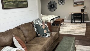 Ein Wohnzimmer mit einem braunen Ledersofa, einer grünen Bank und einem Holztisch mit Stuhl. An den Wänden hängen zwei dekorative Teller und ein gerahmtes Landschaftsbild.