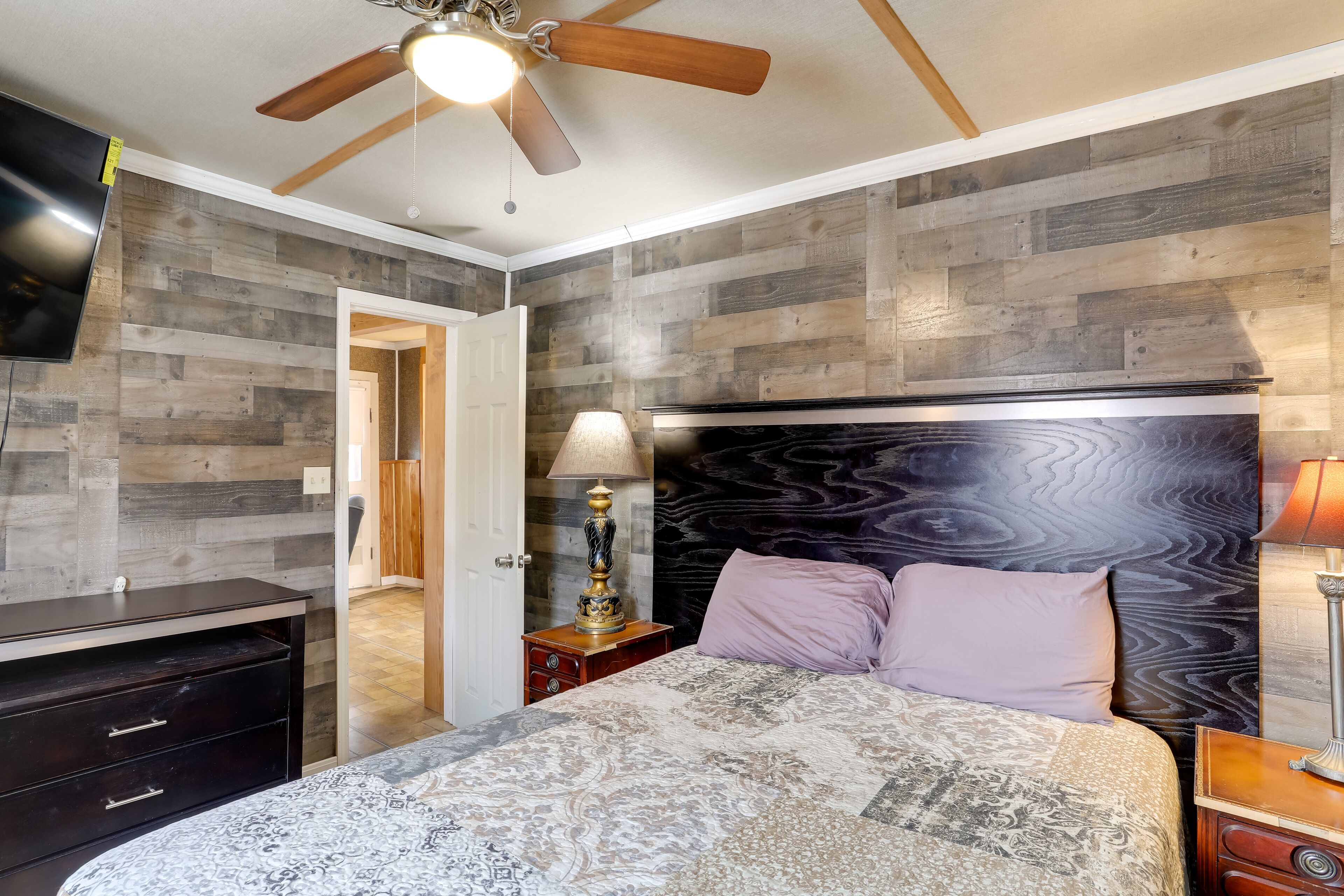 Une chambre à coucher avec une tête de lit en bois, un lit recouvert d’une literie à motifs, une table de chevet avec une lampe et un ventilateur de plafond.