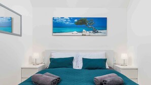 A neatly made bed with a teal bedspread, two pillows, and two rolled towels. There is a framed painting of a beach scene above the bed. - Holiday apartment with panoramic sea views (Klimno)
