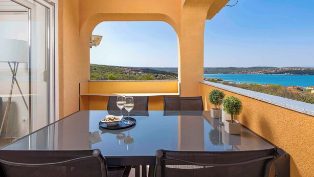 A balcony with a glass table and chairs, a small table with a plate of food and two glasses, and a view of a coastal landscape. - Holiday apartment with panoramic sea views (Klimno)