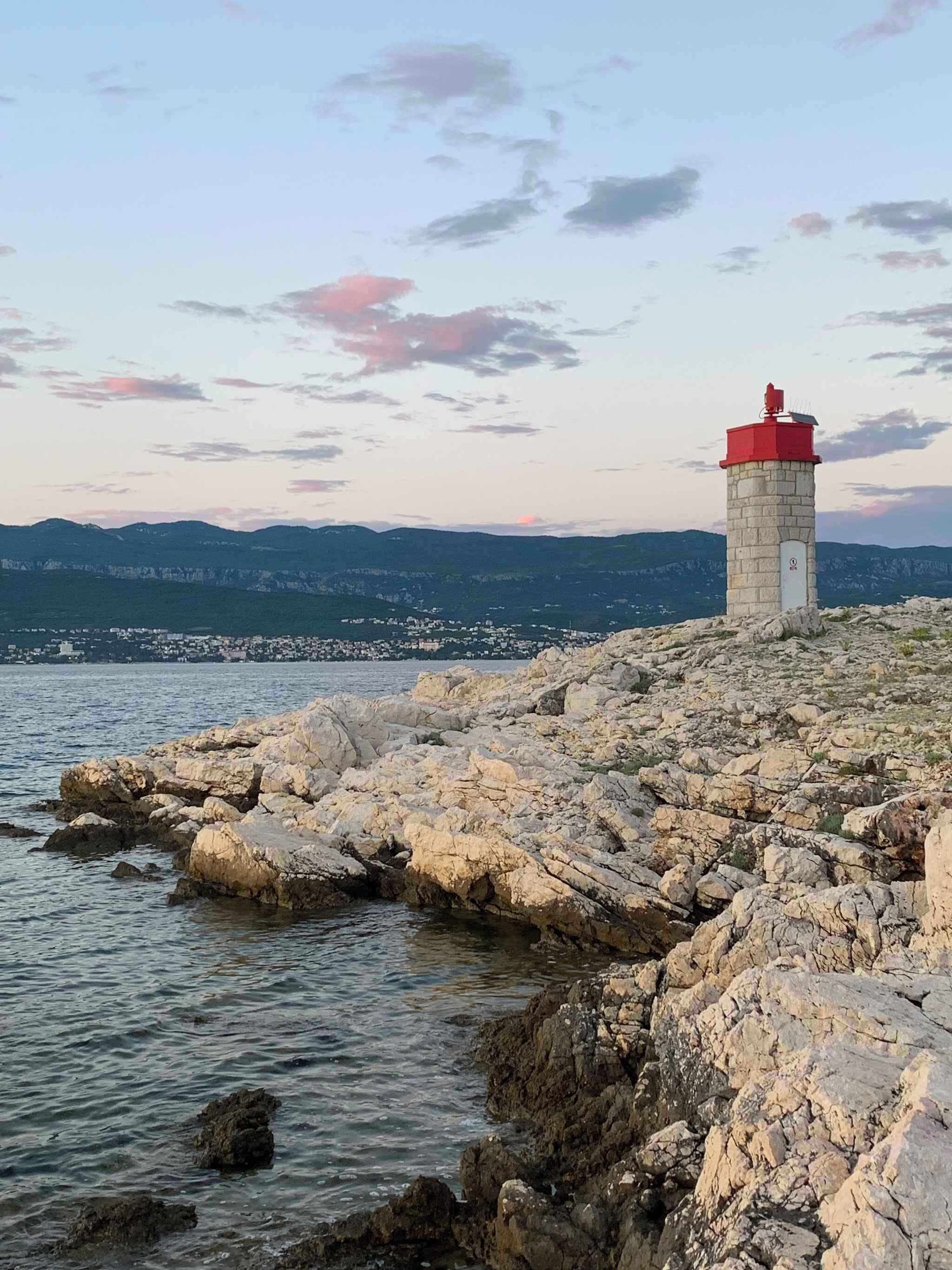 Ein Leuchtturm an einer felsigen Küste mit Blick auf ferne Berge und eine Stadt gegenüber dem Wasser.