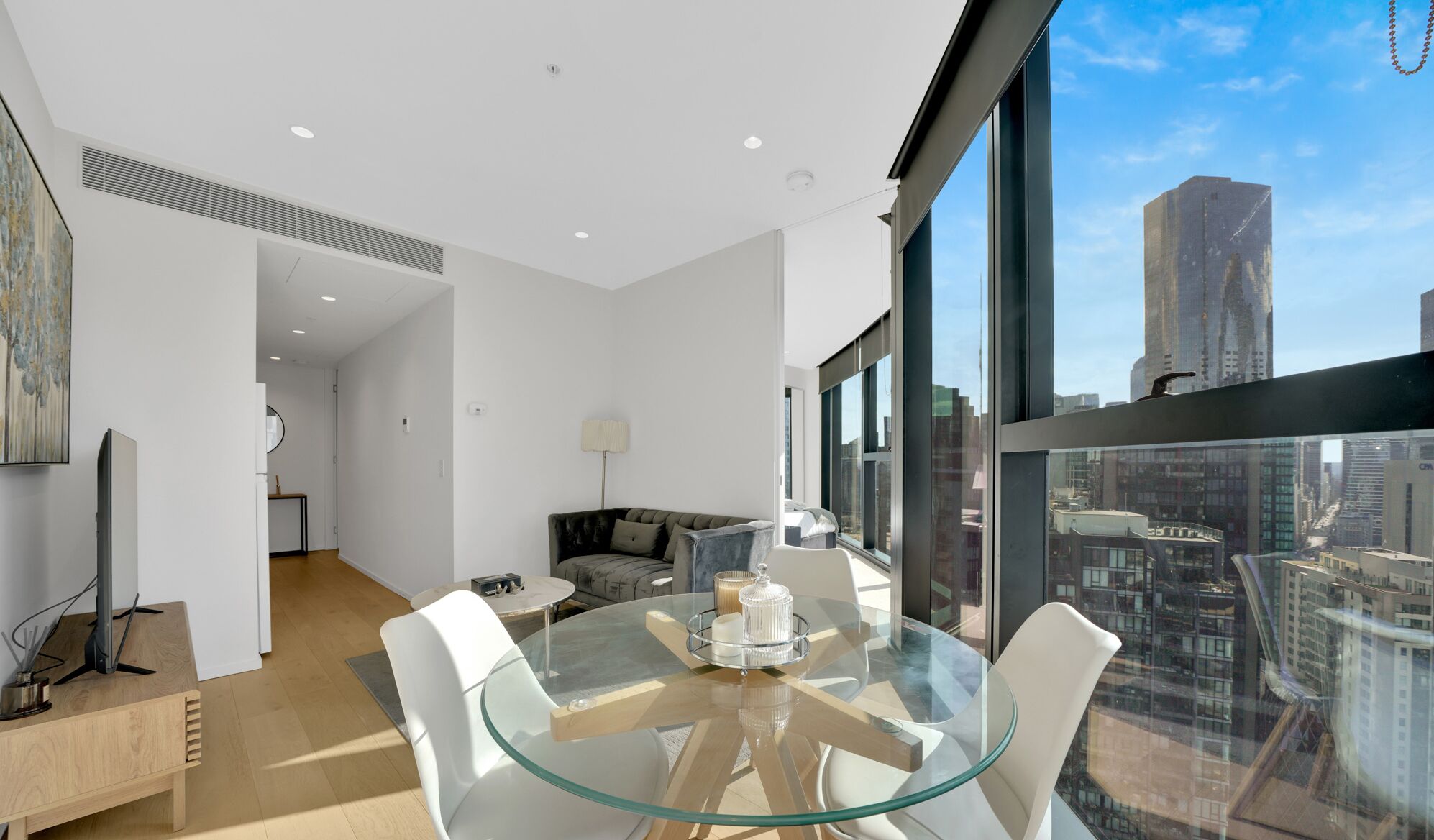 A modern living room with a glass table, white furniture, and a city view through large windows.