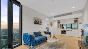 A modern living room with a blue sofa, a small coffee table, and a city view through large windows. - Melbourne Square Apartments (Southbank)