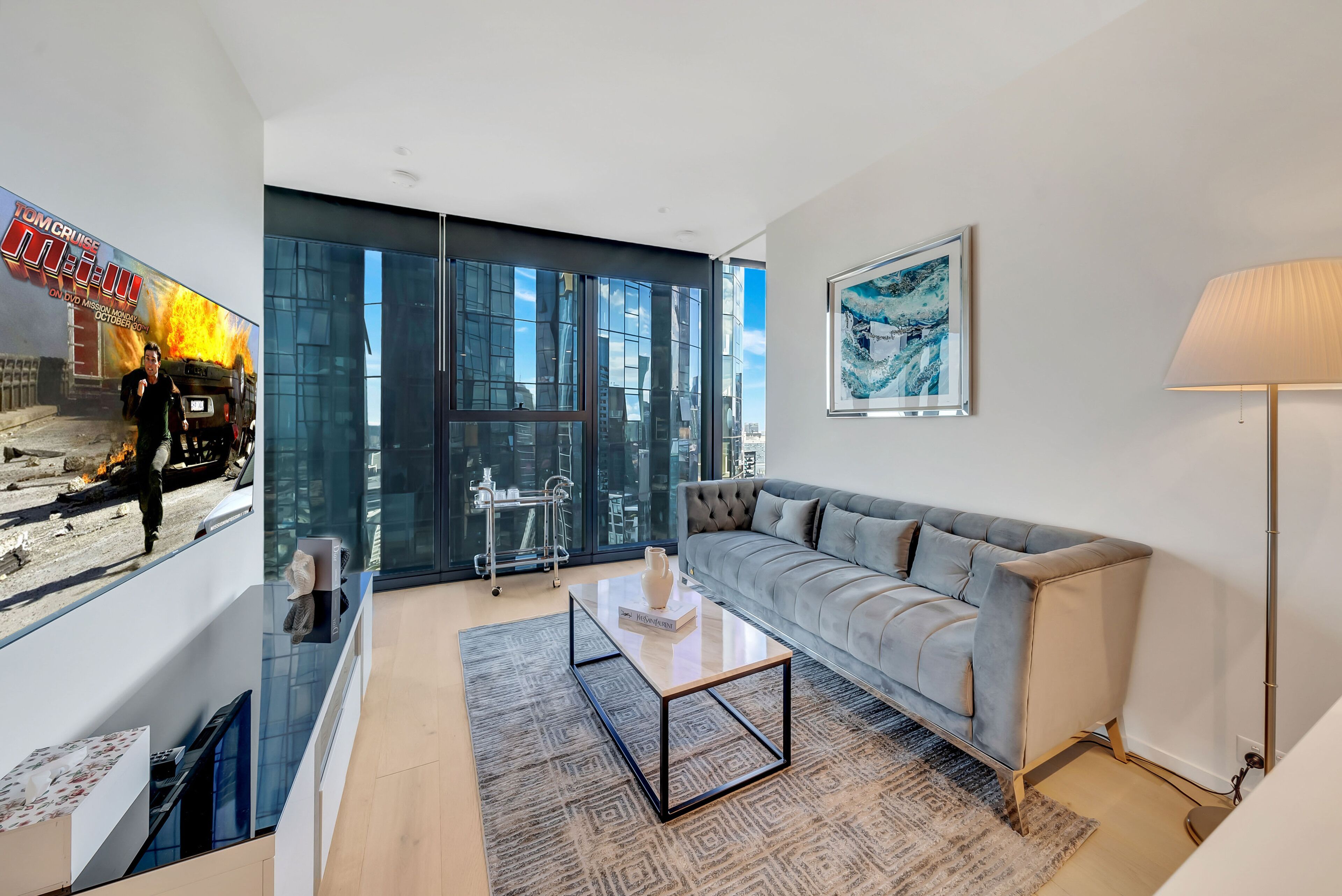 Un salon moderne avec un canapé gris, une table basse en verre et une grande fenêtre donnant sur la ville.
