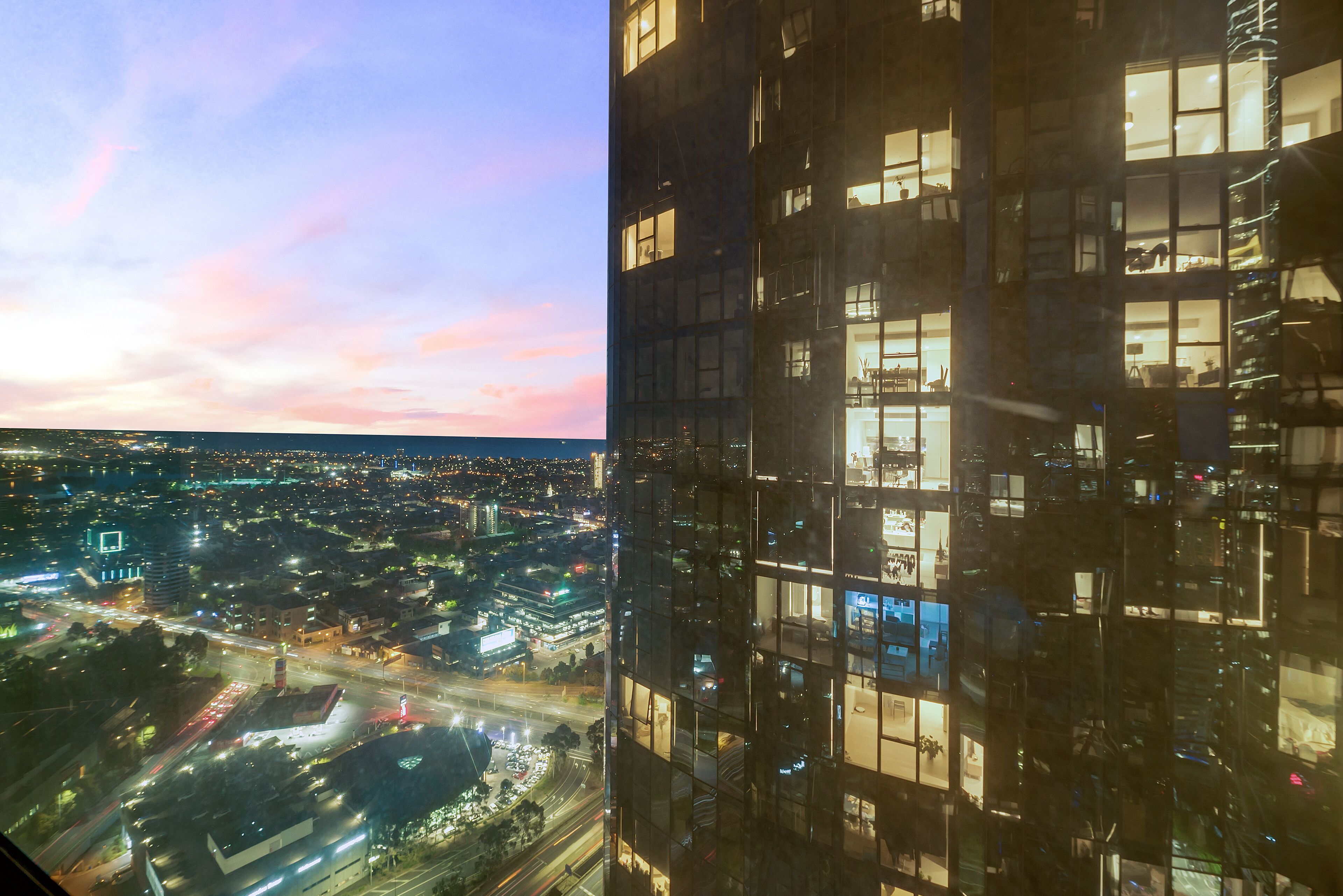 En hög byggnad med många våningar och stora fönster som reflekterar stadens ljus och en solnedgångshimmel.