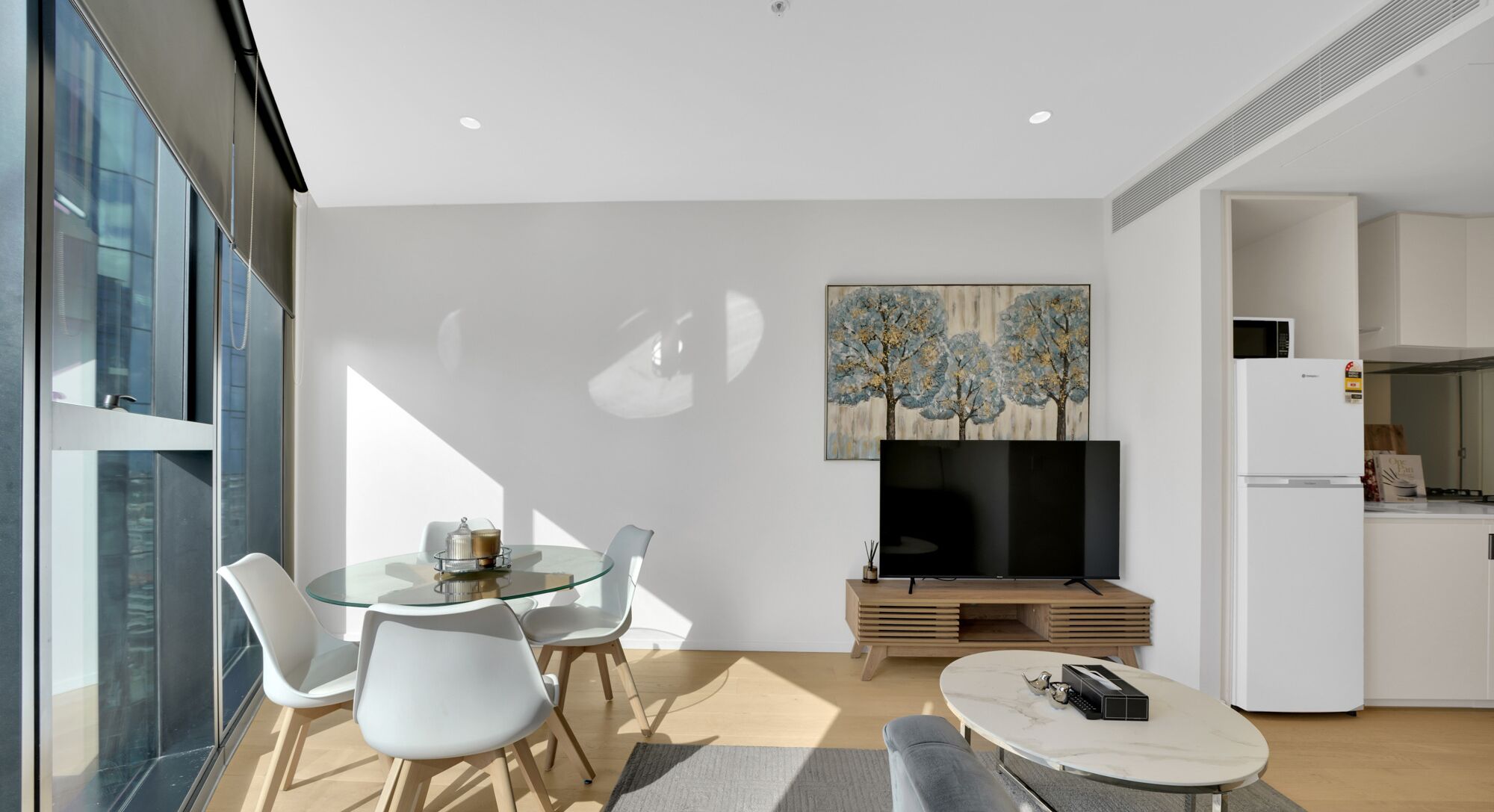 A modern living room with a glass dining table, white chairs, a flat-screen TV, and a painting on the wall.