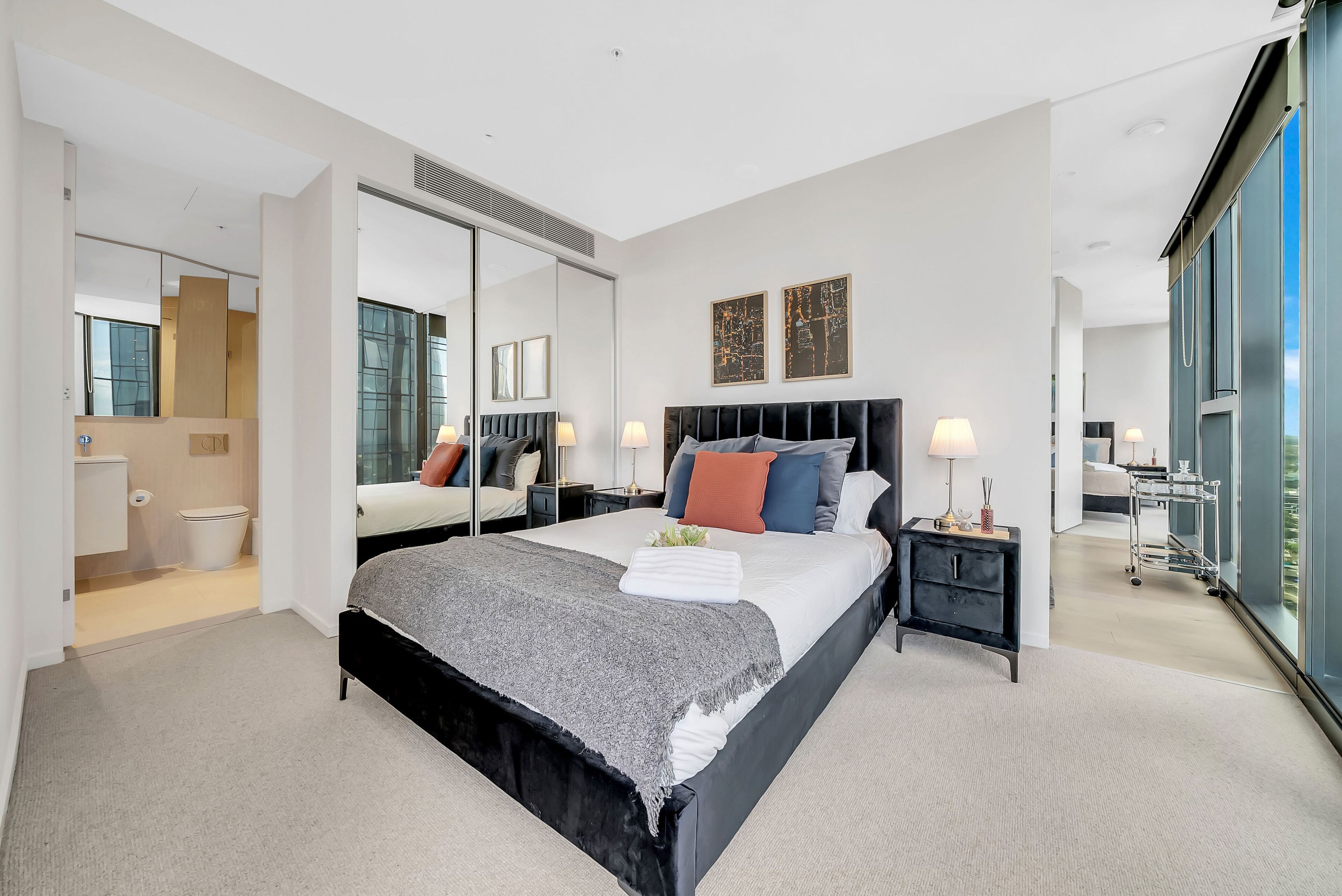 A modern hotel room with a large bed, a nightstand, a mirror, and a view of the city through large windows.