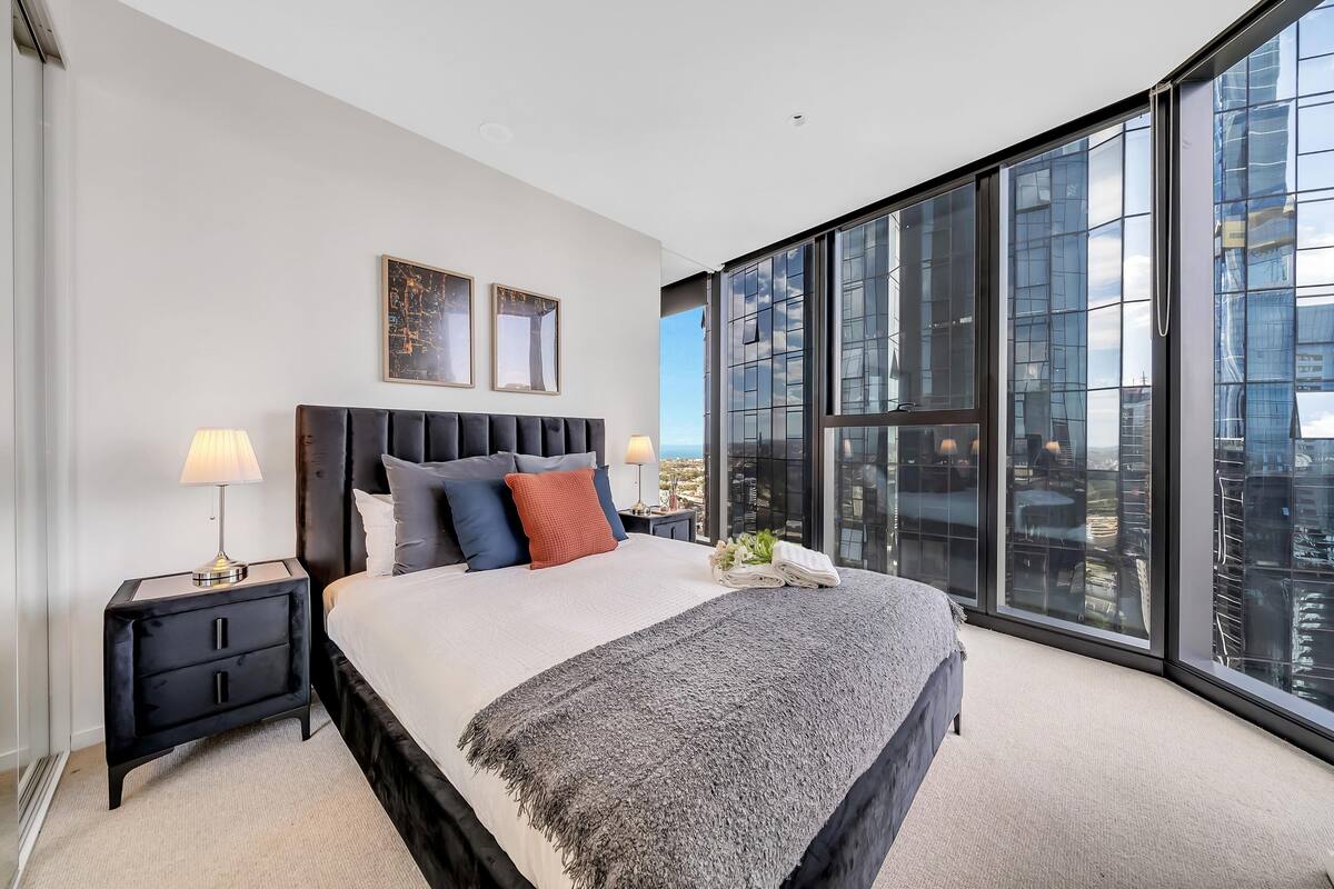 A modern bedroom with a large bed, two bedside tables, and a view of the city through floor-to-ceiling windows.