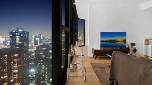 A modern living room with a city view, a large flat-screen TV, and a glass coffee table. - Melbourne Square Apartments (Southbank)