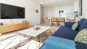 Un salon moderne doté d’un téléviseur à écran plat, d’une console en bois, d’une table basse, d’un canapé bleu et d’un coin repas avec des chaises en bois et une table blanche.