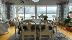 A dining area with a table set for four, surrounded by white chairs, with a view of a lake and trees through large windows. - Awesome home in Värnamo with kitchen (Värnamo)