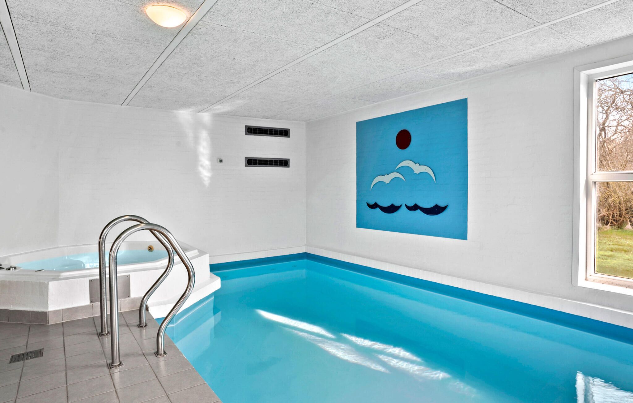 Une piscine intérieure ornée d’une fresque bleue, dotée d’un jacuzzi intégré et d’une fenêtre offrant une vue sur les arbres.