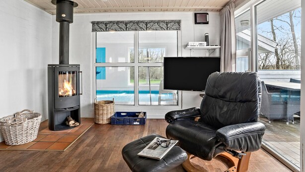 A modern living room with a fireplace, a comfortable leather chair, a television, and a view of a swimming pool. - Nice home in Nørre Nebel with sauna (Nørre Nebel)