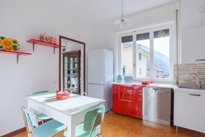Une cuisine compacte avec un réfrigérateur blanc, des placards rouges, un évier et une fenêtre donnant sur des immeubles et des montagnes.