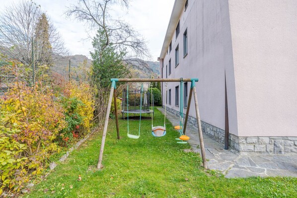Un área con césped, un columpio y un trampolín, rodeada de árboles y un edificio.