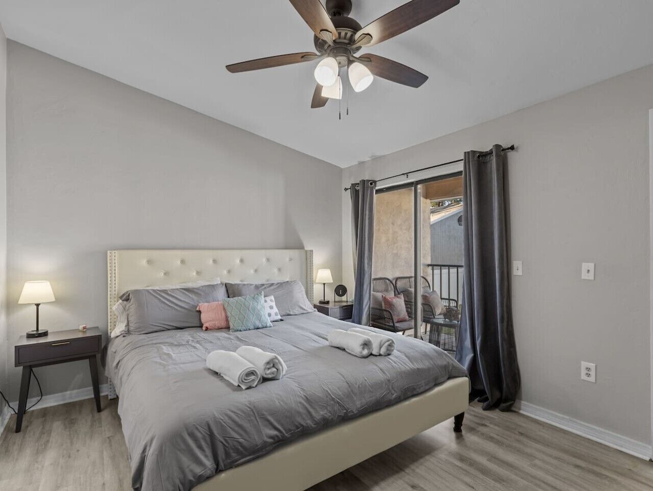 Ein Schlafzimmer mit Bett, Nachttisch, Deckenventilator, Fenster mit Vorhängen und einem Balkon.
