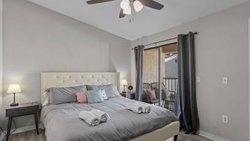 Ein Schlafzimmer mit Bett, Nachttisch, Deckenventilator, Fenster mit Vorhängen und einem Balkon.