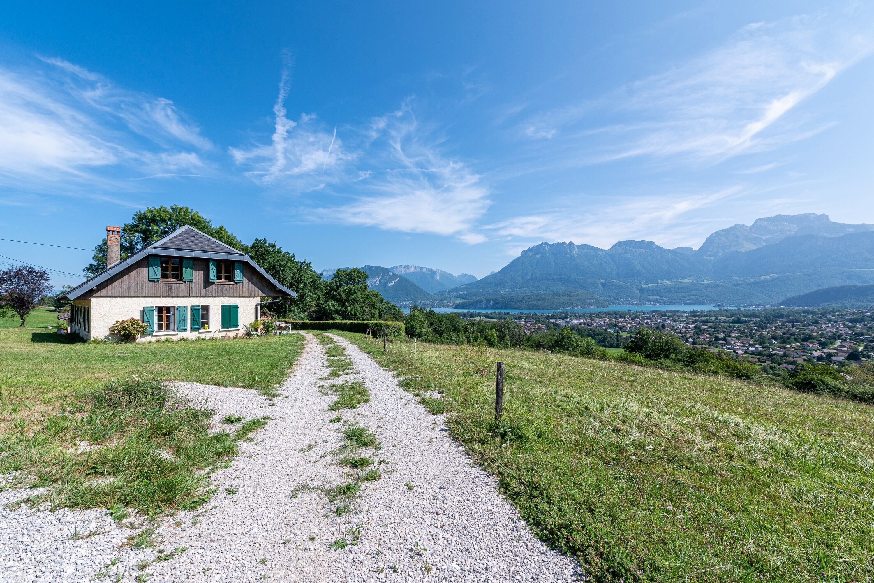 A house with a gravel path leading to it, surrounded by greenery and mountains in the background.