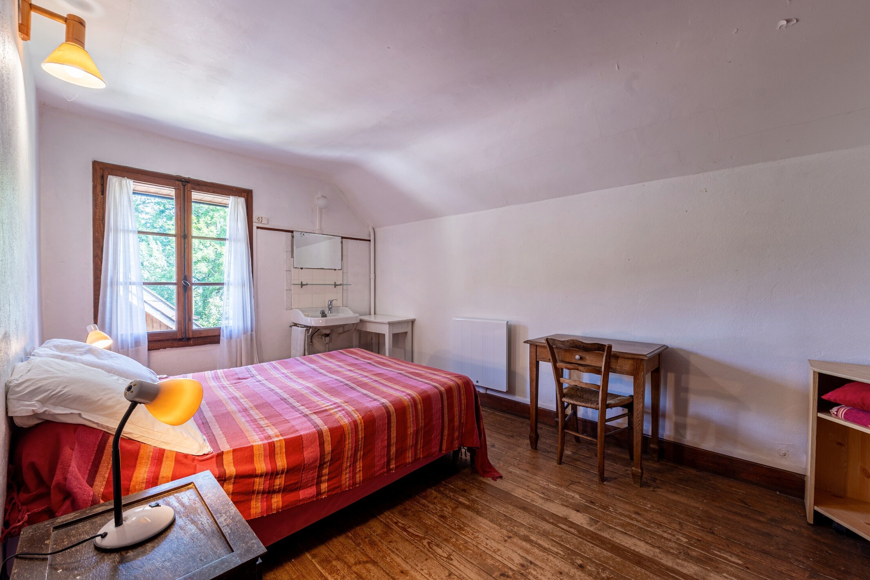 A bedroom with a bed, bedside lamp, wooden desk, chair, and a window with curtains.