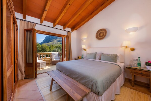 A bedroom with a large bed, wooden ceiling beams, a bench, a bedside table with a lamp, and a view of a balcony with outdoor furniture.