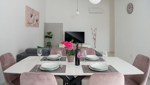 Comedor con una mesa blanca para cuatro personas, rodeada de sillas rosas. Hay una botella de vino y un florero con flores sobre la mesa.