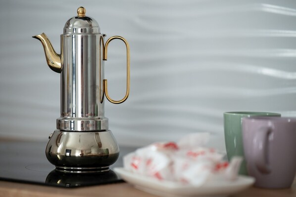 A stainless steel coffee pot with a gold handle and a matching lid, accompanied by a plate of pastries and two mugs.