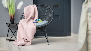Una silla con un cojín y una manta rosa sobre ella, una mesita con un florero de plantas, y una puerta cerrada al fondo.