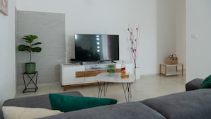 Una sala de estar moderna con un televisor de pantalla plana, un mueble blanco, una mesita con una botella y un vaso, una planta en maceta y un sofá.