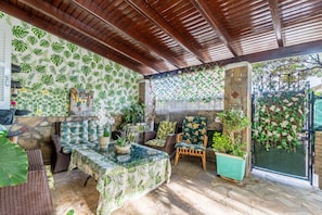 Un patio couvert aménagé avec des meubles en osier, une table en verre et des plantes en pot.