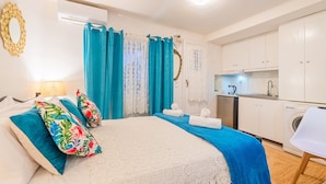 Une chambre à coucher comprenant un lit, des oreillers, une table de chevet, une lampe, une salle de bain avec un rideau de douche, et une kitchenette équipée d’un évier et d’un lave-linge.