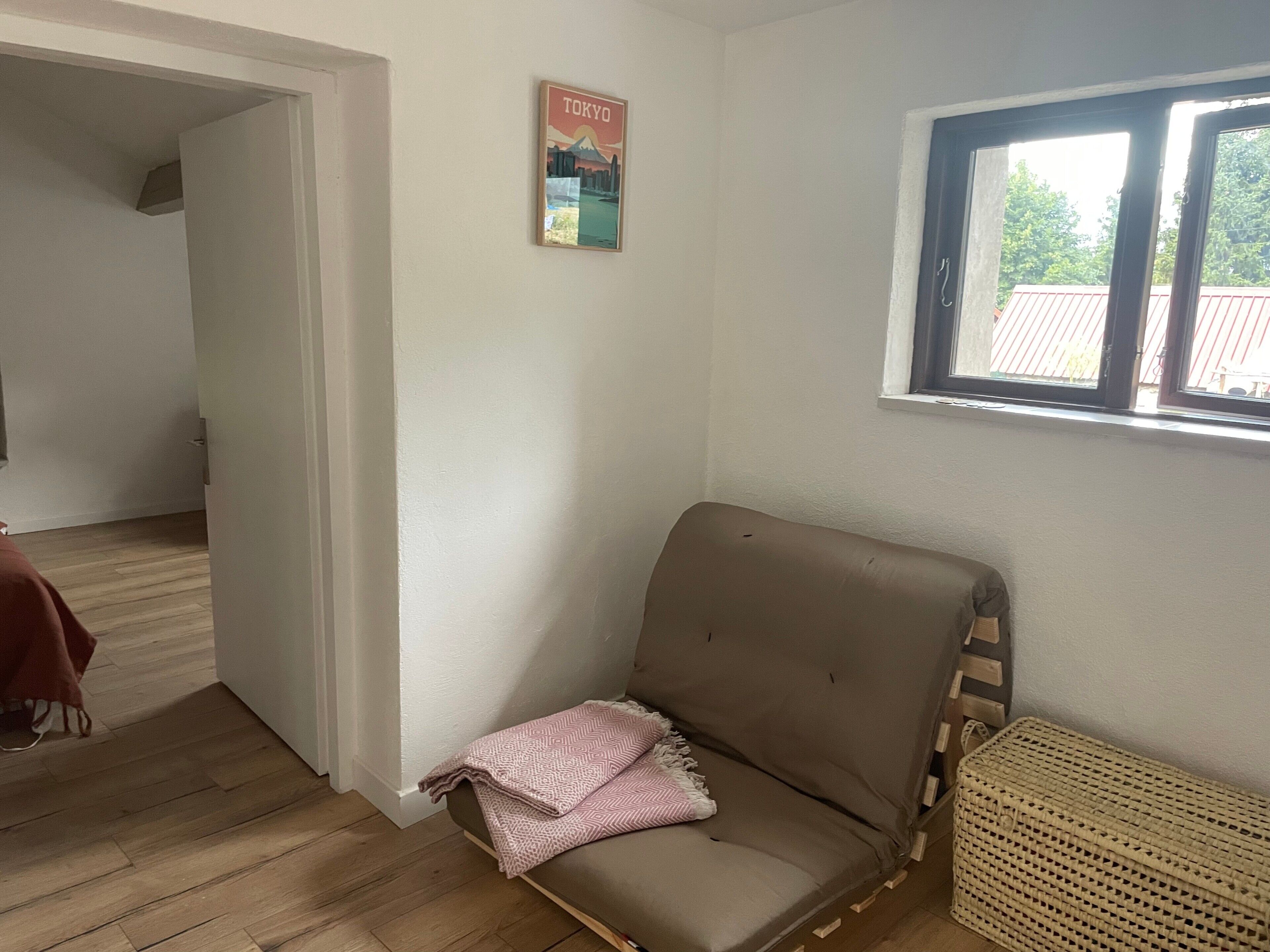 Une pièce avec un canapé marron, un panier en osier, une affiche accrochée au mur et une fenêtre donnant sur des arbres.