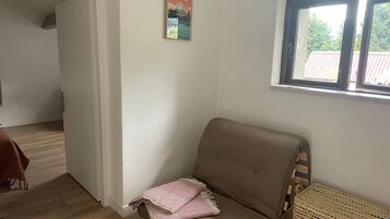 Une pièce avec un canapé marron, un panier en osier, une affiche accrochée au mur et une fenêtre donnant sur des arbres.