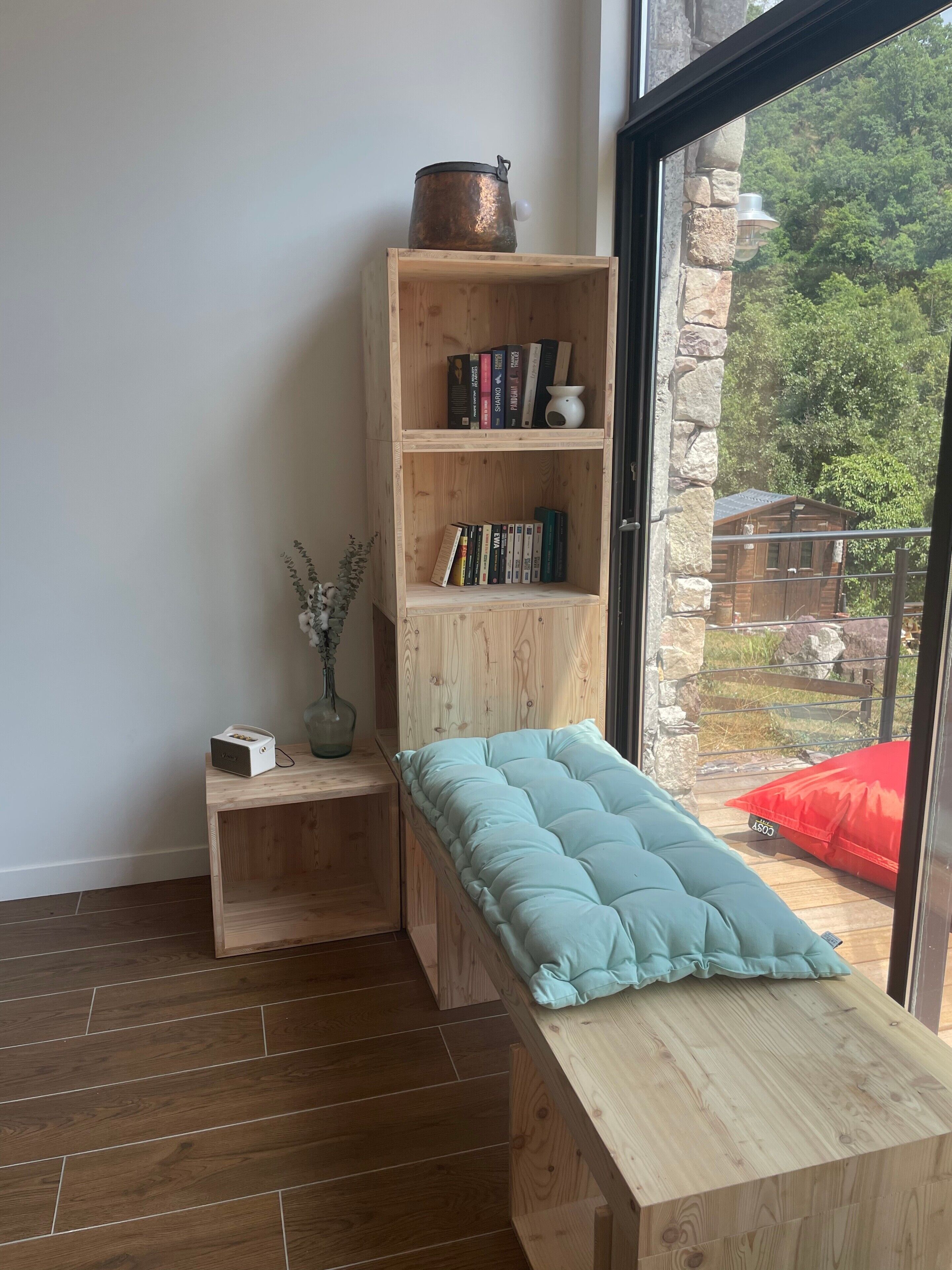 Una habitación con un banco de madera, una estantería con libros y una maceta, y vistas a una cabaña a través de una puerta de cristal.