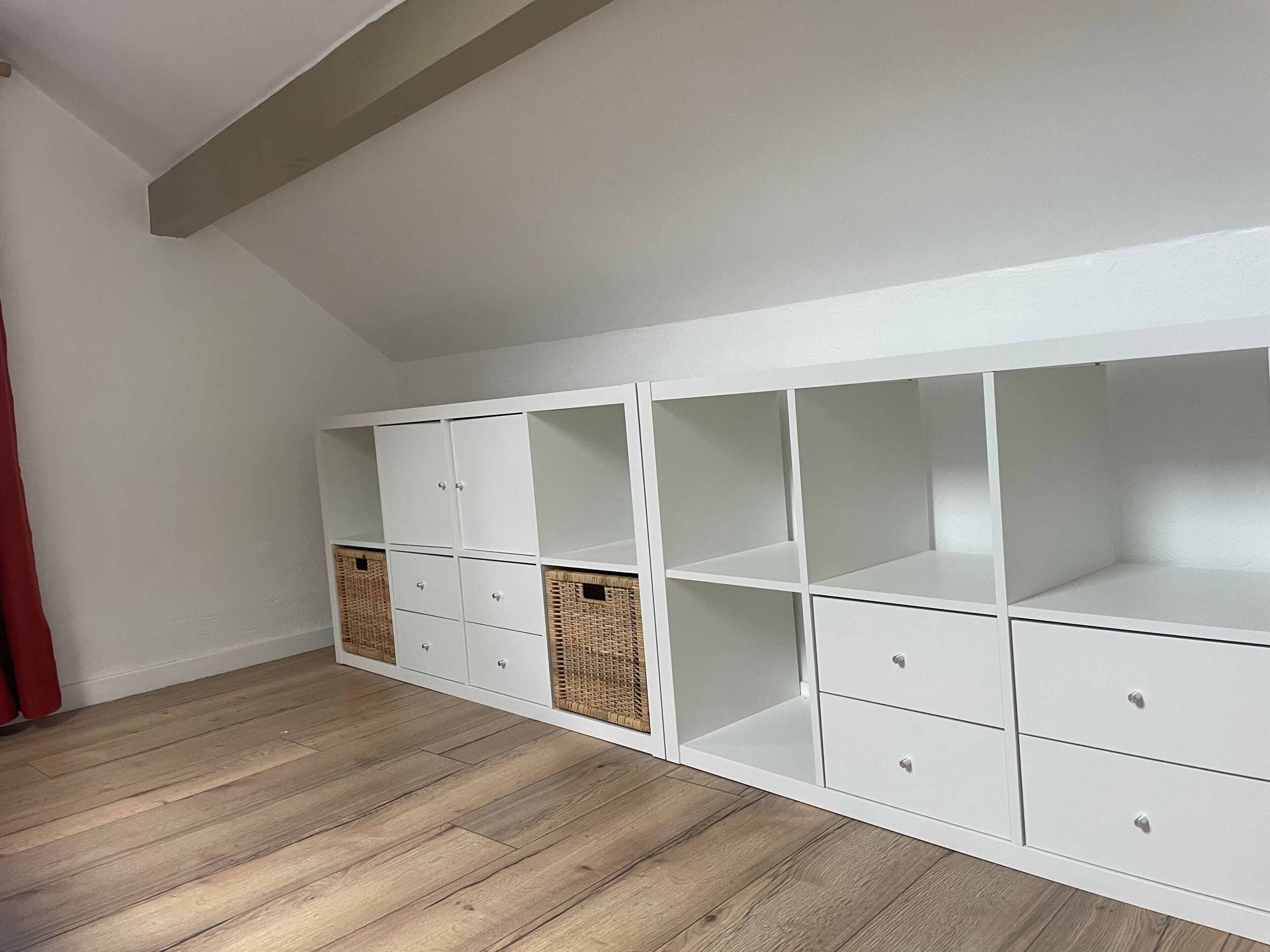 Un módulo de almacenamiento blanco con varios compartimentos y cestas, suelo de madera y techo inclinado.