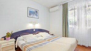 Un cuarto de hotel con una cama, una mesita de noche, un aire acondicionado instalado en la pared, una ventana con cortinas translúcidas y un cuadro enmarcado en la pared.