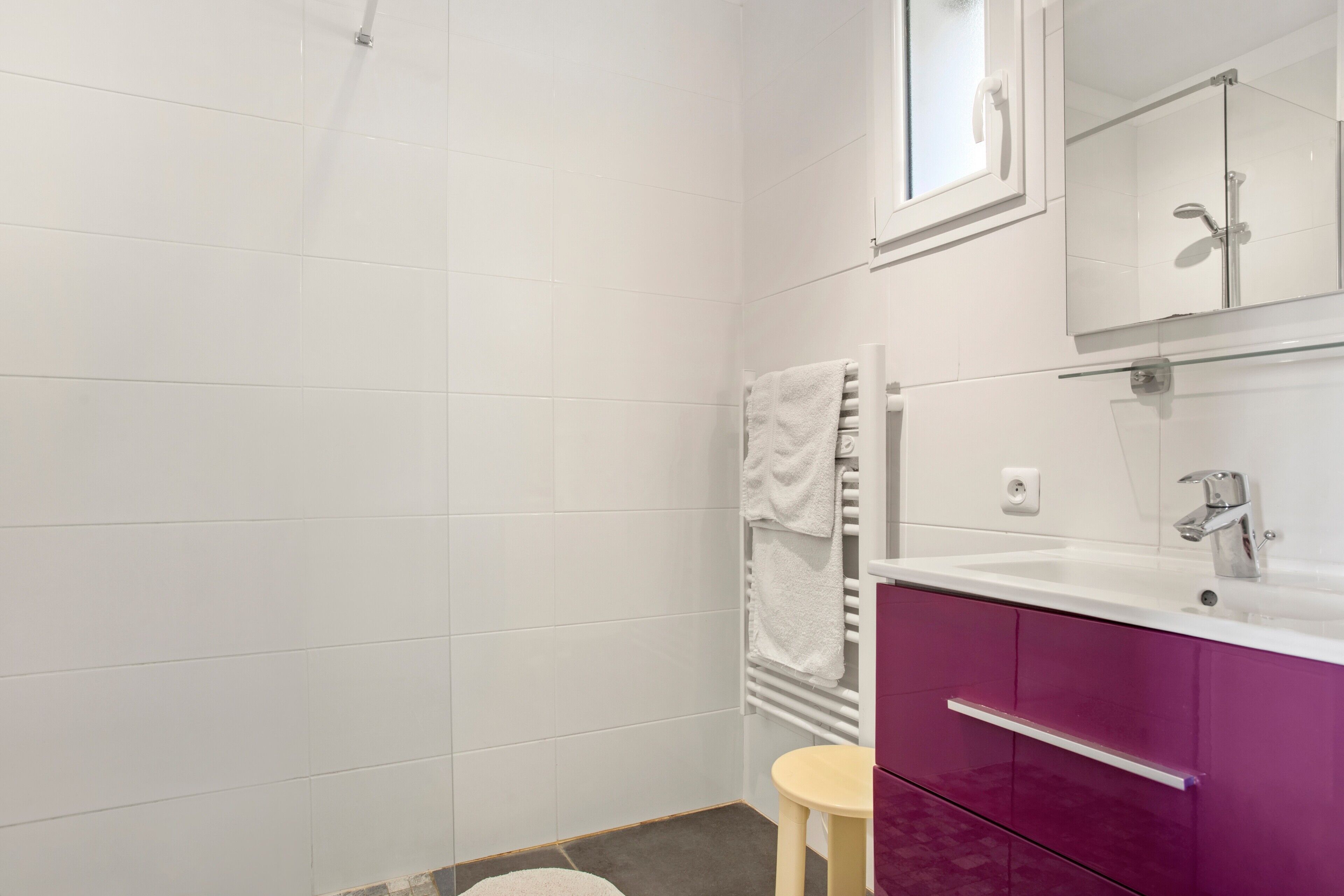 Un baño con azulejos blancos, un tocador morado, un lavabo blanco y un espejo encima.