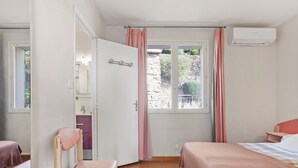 Une chambre à coucher comprenant un lit, une chaise, une petite table, une fenêtre avec des rideaux et un appareil de climatisation.