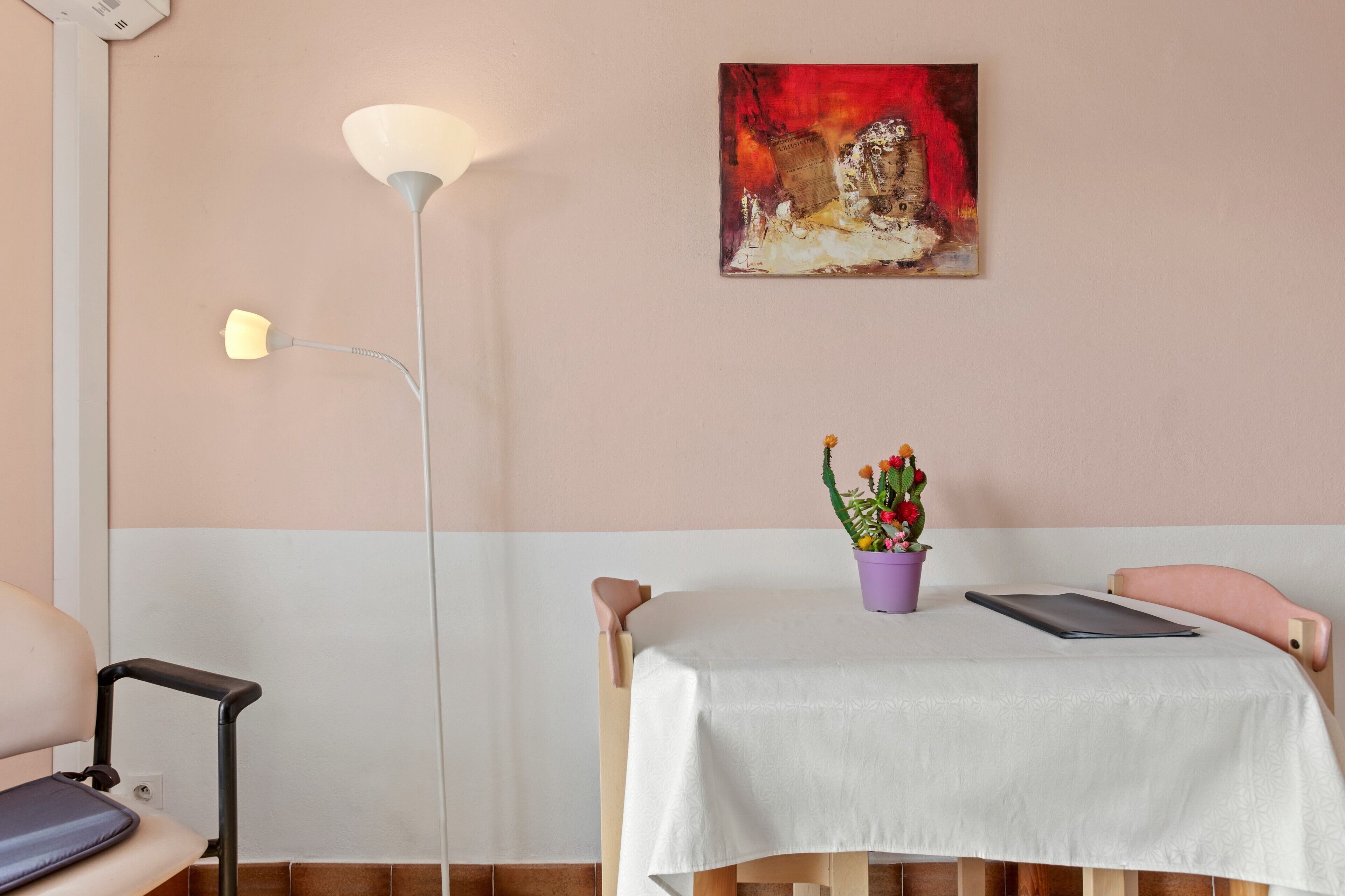 Una habitación con una mesa blanca, una planta morada, un cuadro y una lámpara de pie.
