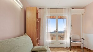 A room with a sofa, a chair, a wooden shelf, a window with curtains, and an air conditioning unit.