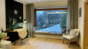 Ett mysigt vardagsrum med ett stort fönster som blickar ut över en snötäckt skog, en bekväm soffa, en fåtölj och en dekorativ vägg med stjärnformade prydnader.