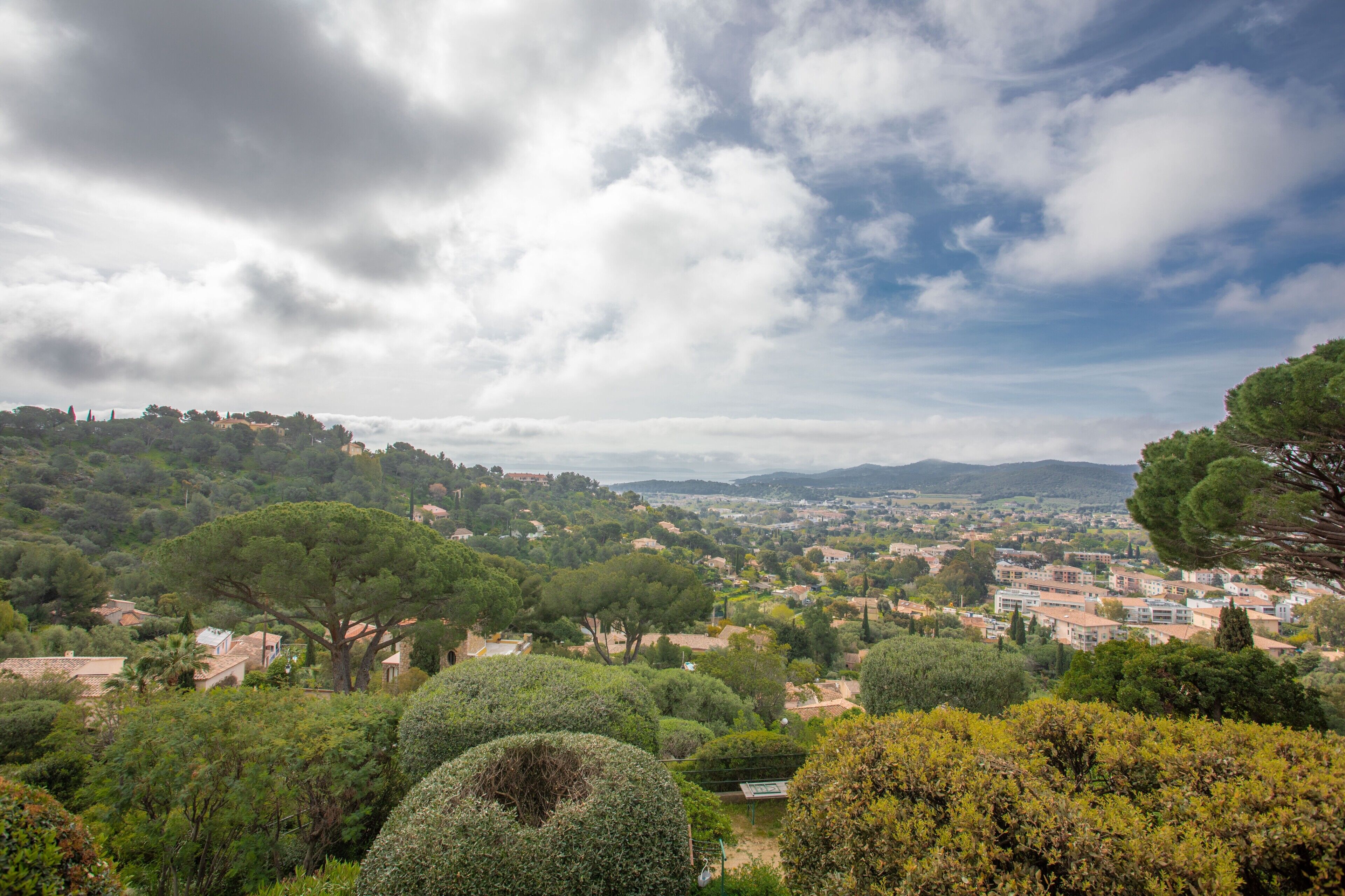 A panoramic view of a town with dense greenery, hills, and a clear sky.