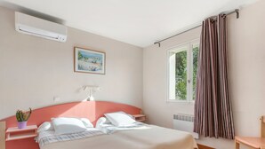 Un dormitorio con cama, mesita de noche, planta en maceta, ventana con cortinas y equipo de aire acondicionado.