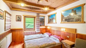Ein Zimmer mit Holzbalkendecke, einem Bett mit zwei Kissen, einem Nachttisch und einem Fenster mit Blick auf Grünfläche.