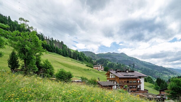 Eine ländliche Berglandschaft mit einem Holthaus und einem kleineren Gebäude im Vordergrund, umgeben von Grünflächen und Hügeln.