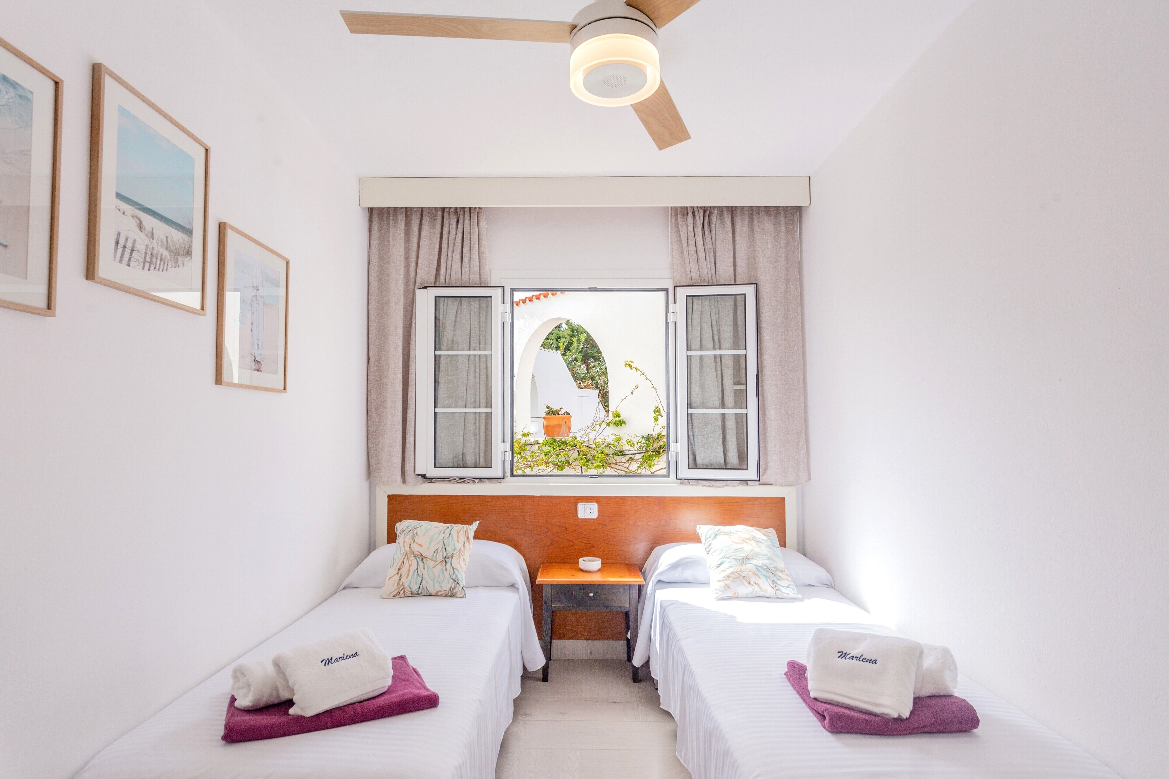 A hotel room with two beds, a ceiling fan, a window with curtains, and framed pictures on the wall.