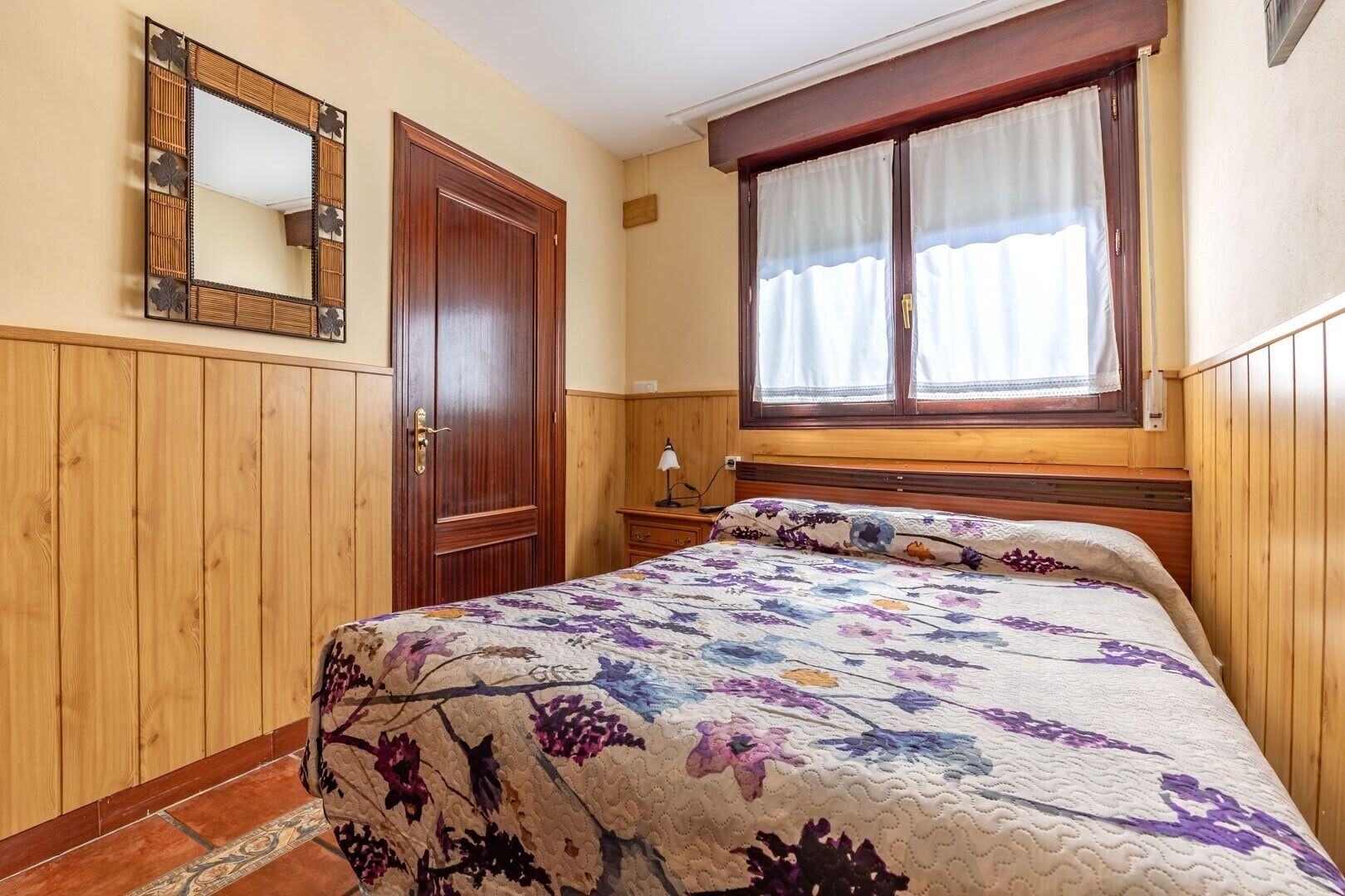 Ein Schlafzimmer mit einem Holzschrank, einem Bett mit Blumendekoration und einem Fenster mit Vorhängen, sowie einem Spiegel an der Wand.