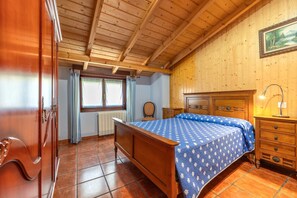 A bedroom with a wooden ceiling, a bed with a blue patterned bedspread, a wooden dresser, and a window with curtains.