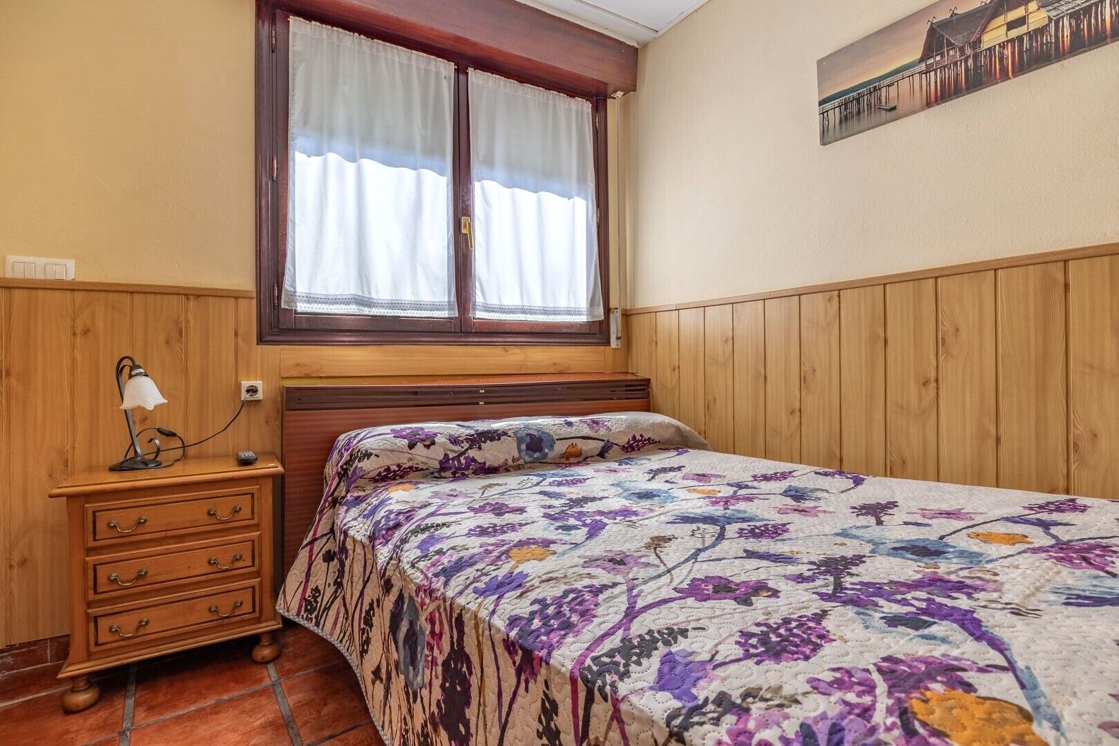 Un dormitori amb llit de fusta, una nitzeta amb làmpada, una finestra amb cortines i un quadre penjat a la paret.