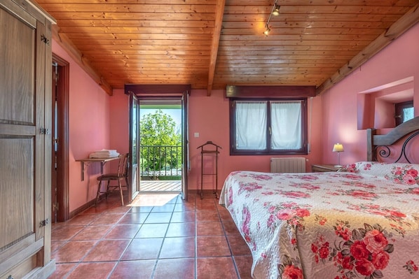 A bedroom with a wooden ceiling, a bed with a floral bedspread, a window with curtains, and a view of greenery outside.