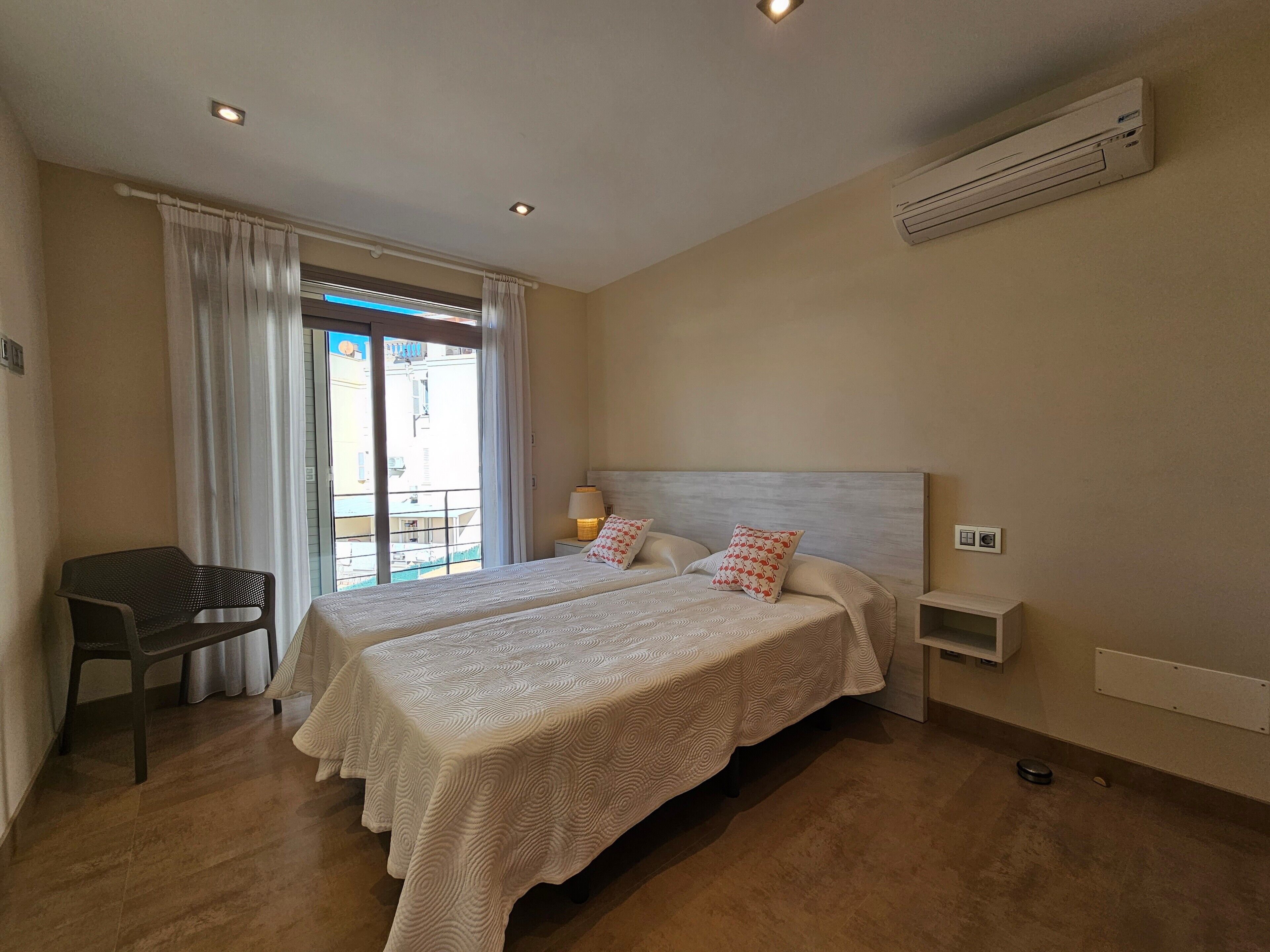 A hotel room with two beds, a chair, a window with curtains, an air conditioning unit, and a view of the outside.